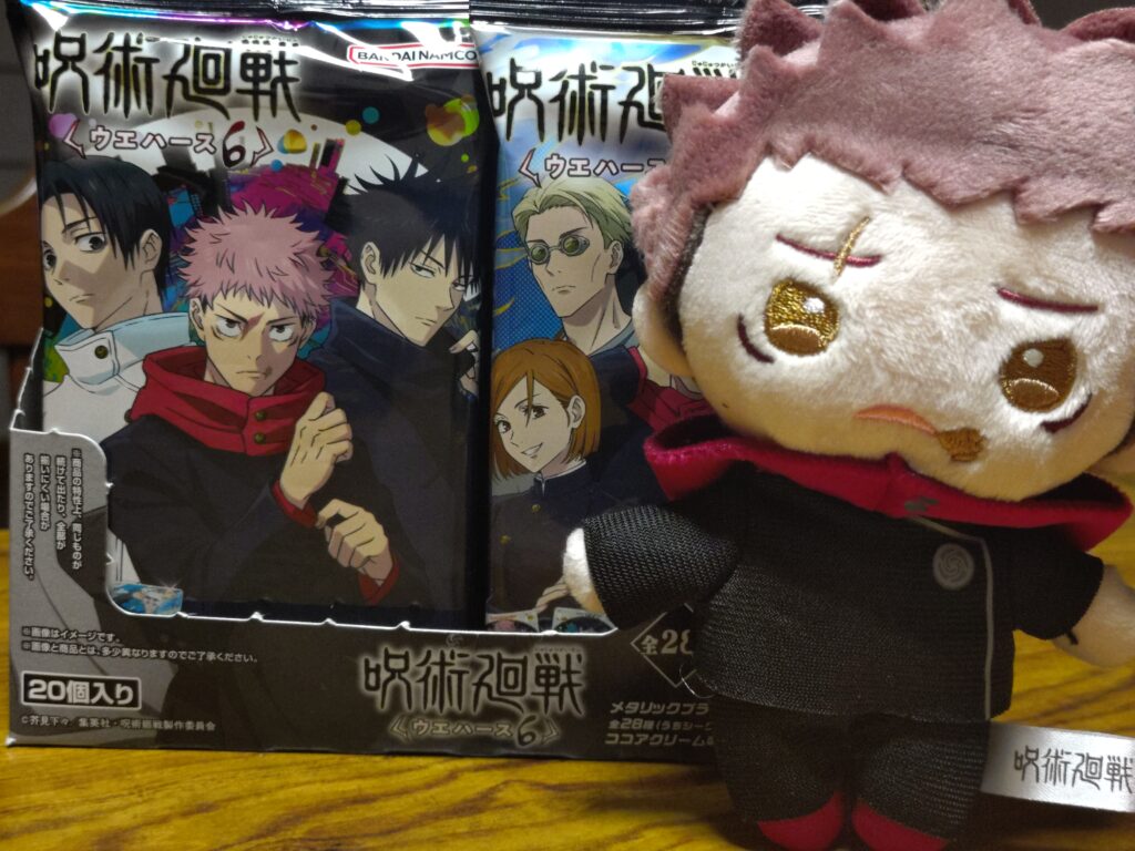 呪術廻戦ウエハース6のパッケージを箱に立てて並べた様子。手前には不安そうな表情の虎杖悠仁ぬいぐるみ。